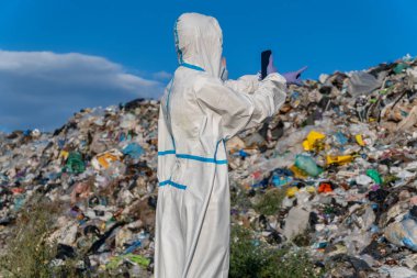 Koruyucu bir takım elbise giyen ve eldiven takan biri, gökyüzünde bulutlar sürüklenirken, çöplük alanındaki büyük bir çöp yığınını işaret ediyor..