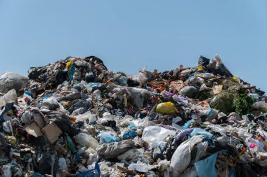 Büyük bir çöp yığını berrak bir gökyüzünün altında, atılan çeşitli materyalleri ve atıkların çevre üzerindeki etkisini sergiliyor..