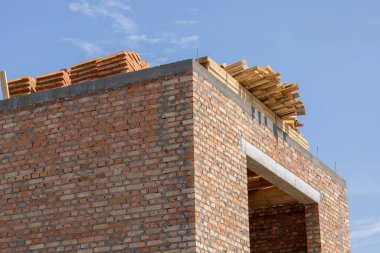 Açık mavi bir gökyüzünün altında, sağlam bir tuğla duvar yükseliyor, düzgünce istiflenmiş ahşap kirişlerle kaplı, bir bina projesinde ilerlemenin sinyallerini veriyor..