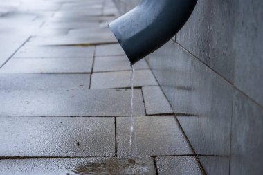 Metal bir drenaj borusundan damlayan su, yağmurlu bir günün huzurlu bir anında, ıslak kaldırımda küçük dalgalanmalar yaratır..