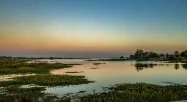 Aşağıdaki sakin su, gökyüzünün renklerini yansıtıyor ve uyumlu bir yansıma yaratıyor. Bitki örtüsü ve uzak ağaçlar manzarayı çerçevelerken, binalar da yakınlardaki bir yerleşim bölgesinde kısmen yapraklarla gizleniyor..