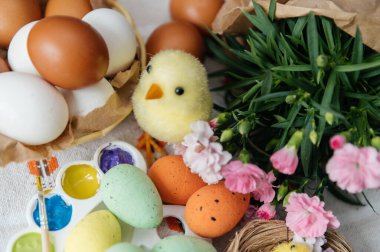 Canlı bir Paskalya yaratıcı düzeneği, doğal ve renkli boyanmış yumurtalar, parlak boyalı bir sanat paleti, ve sevimli sarı piliçler. Pembe karanfiller ve küçük bir yuva bu sanatsal tatil kompozisyonuna şenlikli bir bahar dokunuşu ekler. Üst görünüm.