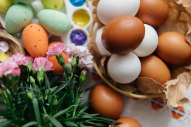 Canlı bir Paskalya yaratıcı düzeneği, doğal ve renkli boyanmış yumurtalar, parlak boyalı bir sanat paleti, ve sevimli sarı piliçler. Pembe karanfiller ve küçük bir yuva bu sanatsal tatil kompozisyonuna şenlikli bir bahar dokunuşu ekler. Üst görünüm.