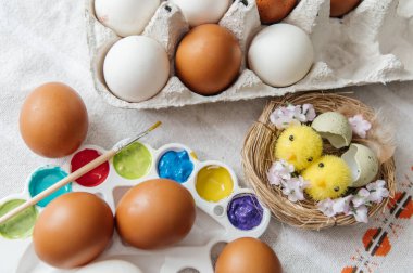 Sanatsal Paskalya 'da doğal kahverengi ve beyaz tavuk yumurtaları, fırçalı canlı bir boya paleti ve yuvada sevimli sarı civcivler bulunur. Pembe karanfiller ve kırsal bir keten arka plan sıcak ve yaratıcı bir bahar atmosferi yaratır. Üst görünüm.