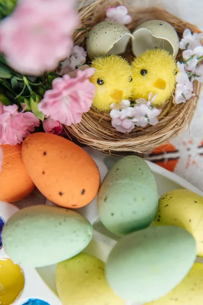 Canlı bir Paskalya yaratıcı düzeneği, doğal ve renkli boyanmış yumurtalar, parlak boyalı bir sanat paleti, ve sevimli sarı piliçler. Pembe karanfiller ve küçük bir yuva bu sanatsal tatil kompozisyonuna şenlikli bir bahar dokunuşu ekler. Üst görünüm.