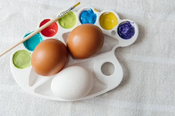 Sanatsal Paskalya 'da doğal kahverengi ve beyaz tavuk yumurtaları, fırçalı canlı bir boya paleti ve yuvada sevimli sarı civcivler bulunur. Pembe karanfiller ve kırsal bir keten arka plan sıcak ve yaratıcı bir bahar atmosferi yaratır. Üst görünüm.