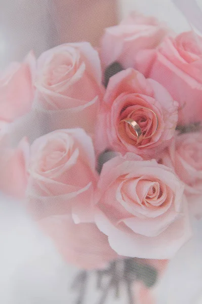 Yumuşak pembe bir gül tomurcuğunun içine yerleştirilmiş iki altın alyans romantik bir yakın plan. Gelin buketi beyaz bir tül peçeyle özenle örtülmüş, rüya gibi, puslu ve zarif bir atmosfer yaratıyor. Düğün fotoğrafçılığı ve gelin dergileri için mükemmel.,