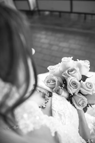 Yüksek açılı siyah-beyaz portre, omuzları açık dantelli gelinlik içinde nefes kesici bir gelin. O, gür bir buket gül tutarken zarif bir şekilde aşağıya bakıyor. Zarif bir gelin mücevheri ve uzun bir peçe klasik görünüşü tamamlıyor..