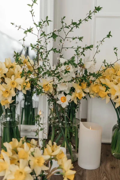 Sarı nergislerin, beyaz narsislerin ve ahşap bir masanın üzerindeki cam vazolarda çiçek açan kiraz dallarının bulunduğu bahar çiçeği kompozisyonu. Paskalya dekorasyonu ve mevsimsel uyanış..