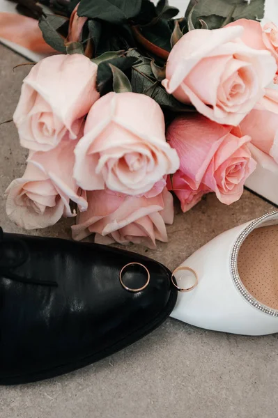 Siyah deri bir damat ayakkabısına takılmış altın alyansların ve beyaz süslemeli gelinlik topuğunun olduğu romantik bir düğün. Taze pembe güllerden oluşan güzel bir buket, arka planda, taş bir yüzeyde yatıyor..