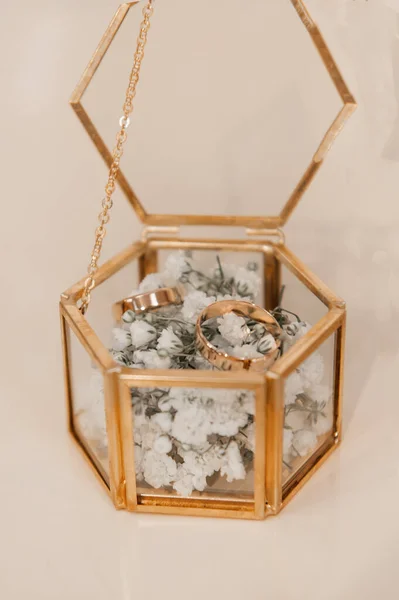 Bir çift zarif altın alyans, pirinç çerçeveli klasik tarzda altıgen bir mücevher kutusunun içinde narin bir beyaz çigila (bebek nefesi) üzerinde dinleniyor. Bu yumuşak, romantik yakın plan, evlilik için mükemmel, zamansız bir düğün detayını yakalar.