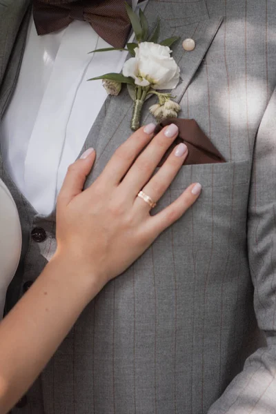 Gri çizgili takım elbise, kahverengi papyon ve beyaz gül yaka çiçeği giyen bir damadın dikey yakın çekimi. Altın bir alyans ve yumuşak pembe bir manikür içeren gelinin eli nazikçe göğsüne dayanıyor. Zarif ve klasik düğün töreni detayları