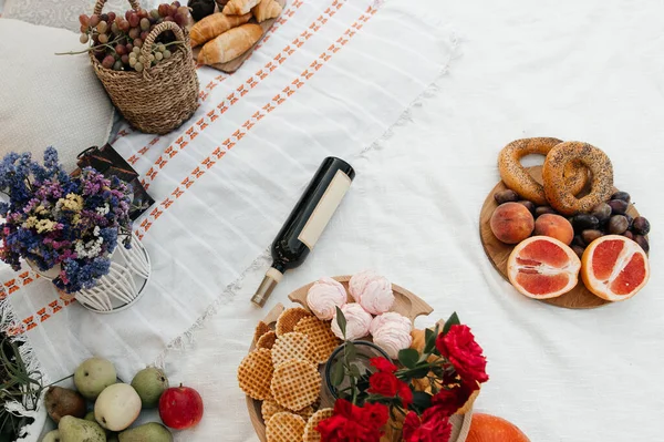 Beyaz desenli bir battaniyenin üzerinde güzel düzenlenmiş bir pikniğin yatay yüksek açılı görüntüsü. Kurulum bir şişe şarap, bir sepet üzüm, kruvasan ve simit içeriyor.