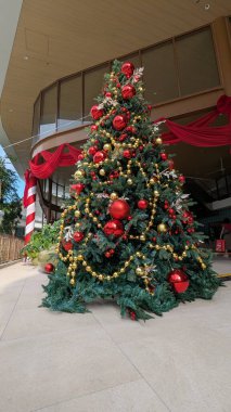 Kırmızı ve altın süslerle süslenmiş kapalı Noel ağacı, boncuk çelenkler ve gümüş yapraklar, bayram temalı bir alanda şenlikli kırmızı kumaşa karşı hazırlanır..