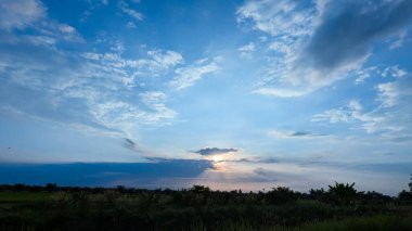 Parlayan bulutlar ve siluetli ağaçlarla gün doğumu veya günbatımı gökyüzü, sakin ve doğal bir manzara görüntüsü yakalıyor..