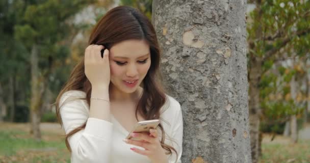 Aantrekkelijke Japans meisje dragen witte jurk texting onder boom en glimlachen — Stockvideo