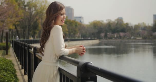 Menina japonesa atraente vestindo vestido branco assistindo as flores de cerejeira no parque sorri para o médio camrea deslizante tiro 4K — Vídeo de Stock