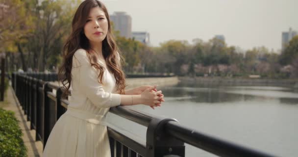 Piękne młode kobiety japoński pochyla się agaisnt poręczy w parku uśmiechy na aparat 4k — Wideo stockowe