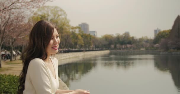 Beautiful Japanese young women wearing white watches the water and cherry blossoms smiles at the camera — Stock Video