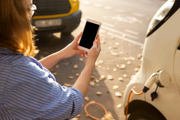 Mani con smartphone su uno sfondo di noleggio auto presso la stazione di ricarica — Foto Stock