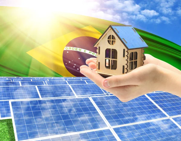 The photo with solar panels and a woman\'s palm holding a toy house shows the flag of Brazil in the sun.