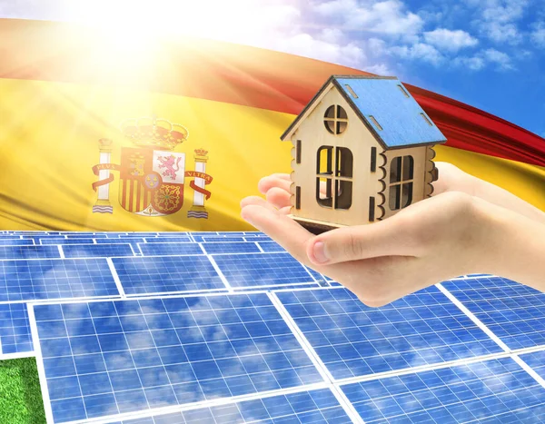 The photo with solar panels and a woman\'s palm holding a toy house shows the flag of Spain in the sun.