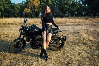 Siyah elbiseli güzel kız motosiklete biner ve kameraya poz verir. Akşam batımı