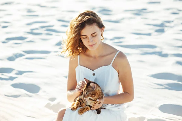 Linda chica en vestido blanco en la playa abraza a su gato de bengala — Foto de Stock