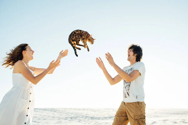 Alegre joven pareja divirtiéndose en la playa con su bengala gato — Foto de Stock
