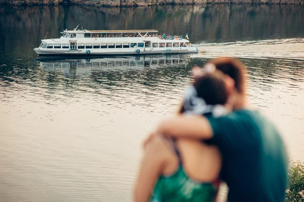 Lovers sitting on the bank of river against background of ship — Stock Photo, Image