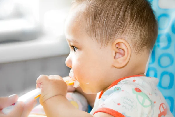 Small happy child sitting in a chair and eats yogurt from whose face is marred in baby food — Stock Photo, Image