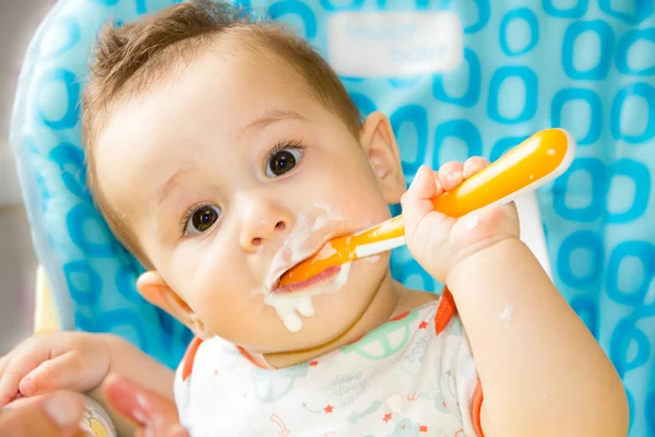 Small happy child sitting in a chair and eats yogurt from whose face is marred in baby food — Stock Photo, Image