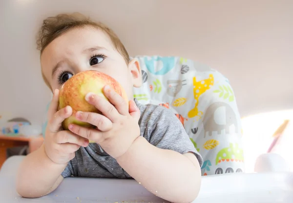 Small happy child sitting in a chair and eats Apple from whose face is marred in baby food, the concept of family, child health, feed the baby, eat at home, funny expression face — Stock Photo, Image