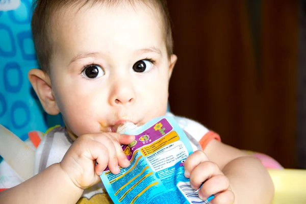 Niño feliz bebé sentado en la silla y come comida de un tubo por sí mismo, el niño estaba sosteniendo paquete de puré de frutas, primer plano, en la cocina, empezar a alimentar — Foto de Stock