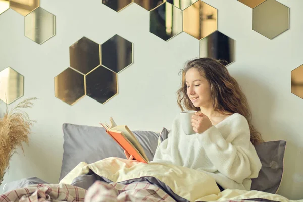 Vrouw zit in bed en drinkt koffie. genieten van de vroege ochtend met een boek in de hand. gezellige winterochtend. — Stockfoto
