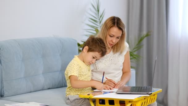 Fiatal anya vagy dada segít egy kisfiúnak befejezni egy feladatot egy online iskolában. Anya és fia együtt csinálják a házi feladatot. — Stock videók