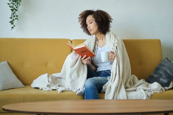 Красивая молодая афроамериканка читает книгу, удобно лежащую на спине на диване в гостиной в джинсах. — стоковое фото