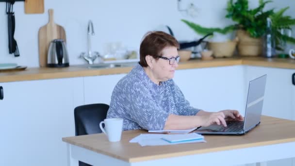 Mujer mayor que utiliza el ordenador portátil para navegar por Internet en su cocina. El concepto de empleo de alto nivel, seguridad social. Señora madura sentada en el trabajo escribiendo una computadora portátil en una oficina en casa. Vídeo filmado — Vídeos de Stock