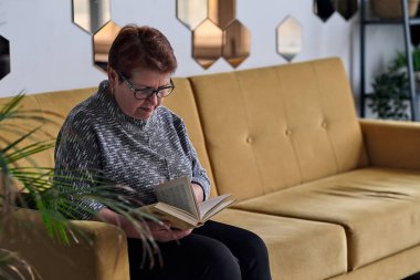 Evdeki kanepede günlük giysiler içinde kitap okuyan güzel yaşlı bir kadın. Gözlüklü, iyi göremeyen son sınıf öğrencisi bir kadın bir kitaba bakıyor..