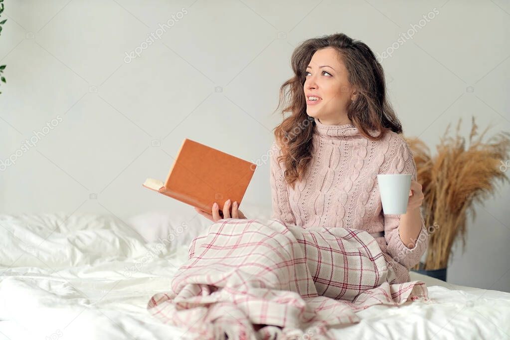 Young woman at home sitting near window relaxing in her living room reading book and drinking coffee or tea.