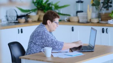 Üst düzey kadın mutfağında web sörfü yapmak için dizüstü bilgisayar kullanıyor. Kıdemli iş ve sosyal güvenlik kavramı. Olgun bir kadın iş yerinde oturup bir ofiste defter bilgisayarı yazıyor. Çekilen video