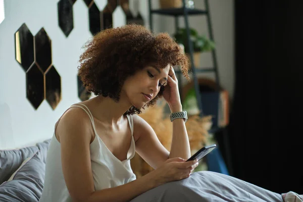 Black woman smiling and using smartphone on bed