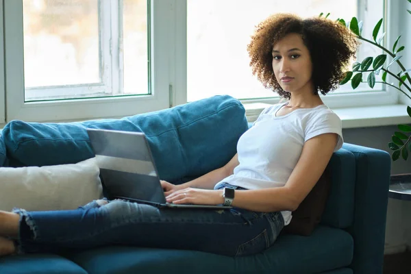 Az afro-amerikai nő otthon laptopot használ. Gyönyörű fiatal hölgy ül a kanapén és egy laptopon dolgozik. szabadúszó vagy távoktatási koncepció — Stock Fotó
