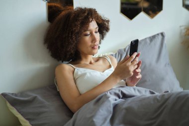 Sakin ol, pijamalı güzel Afrikalı kadın tek başına yatakta yatarken akıllı telefon kullanıyor..
