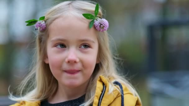 Πορτρέτο ενός υπέροχου κοριτσιού, τριφύλλι λουλούδι στα μαλλιά. χαριτωμένο κορίτσι 5 ετών πονηρά μάτια κοιτάζοντας μακριά με ένα χαμόγελο ματιά στην κάμερα. Κλείστε Shot βίντεο. Πλάνα αργής κίνησης — Αρχείο Βίντεο
