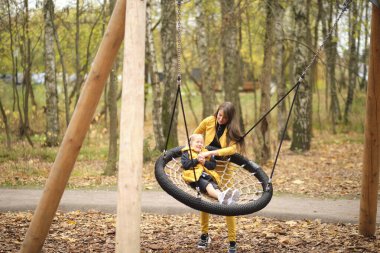 Genç kadın küçük kızını salıncakta sallıyor. Anne ve kızı birlikte eğleniyorlar. sonbahar modası