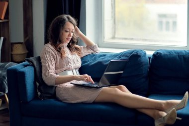 Evdeki güzel esmer kadın kanepeye ya da kanepeye oturmuş dizüstü bilgisayarını kullanıyor ve gülümsüyor.