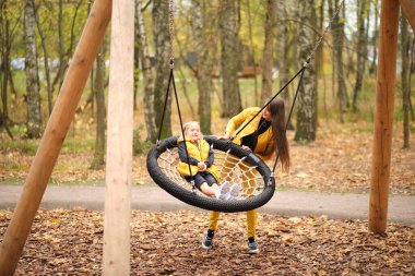 Genç kadın küçük kızını salıncakta sallıyor. Anne ve kızı birlikte eğleniyorlar. sonbahar modası