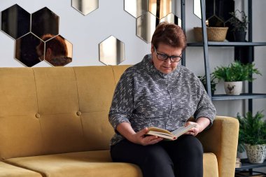 Evdeki kanepede günlük giysiler içinde kitap okuyan güzel yaşlı bir kadın. Gözlüklü, iyi göremeyen son sınıf öğrencisi bir kadın bir kitaba bakıyor..