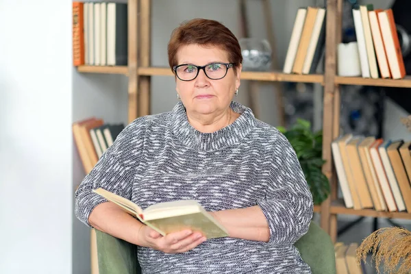 Belle vieille femme en vêtements décontractés lisant sur le canapé à la maison. — Photo